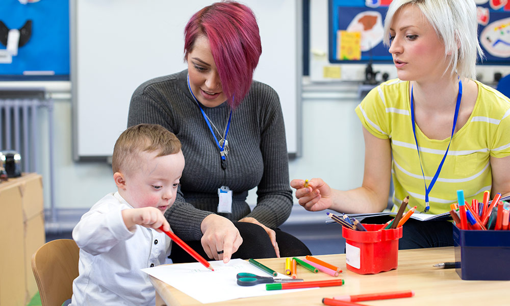 Nursery teachers sitting with young boy with Down Syndrome who is drawing.